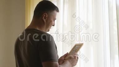一个身穿卡其色t恤的孤独男子站在窗边，手里拿着一张木架上的照片.. 男人看着照片，